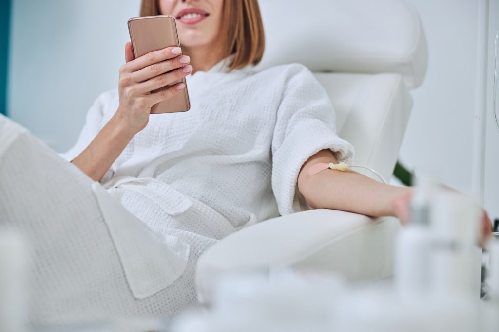 Woman in hospital looking at mobile phone while needle is in arm, Vitamin Therapy Iv Drip Infusion In Women Blood | Bellava Aesthetics in Crestview Hills, KY